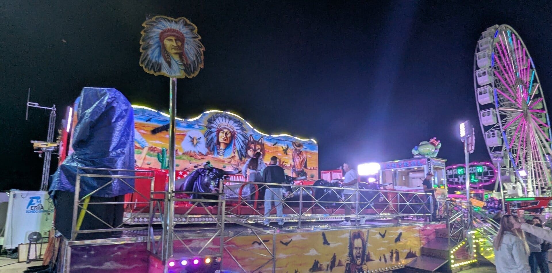 Atracción de toros y caballos mecánicos con decorado del Oeste en la feria de Dénia, con un jefe indio pintado en el fondo