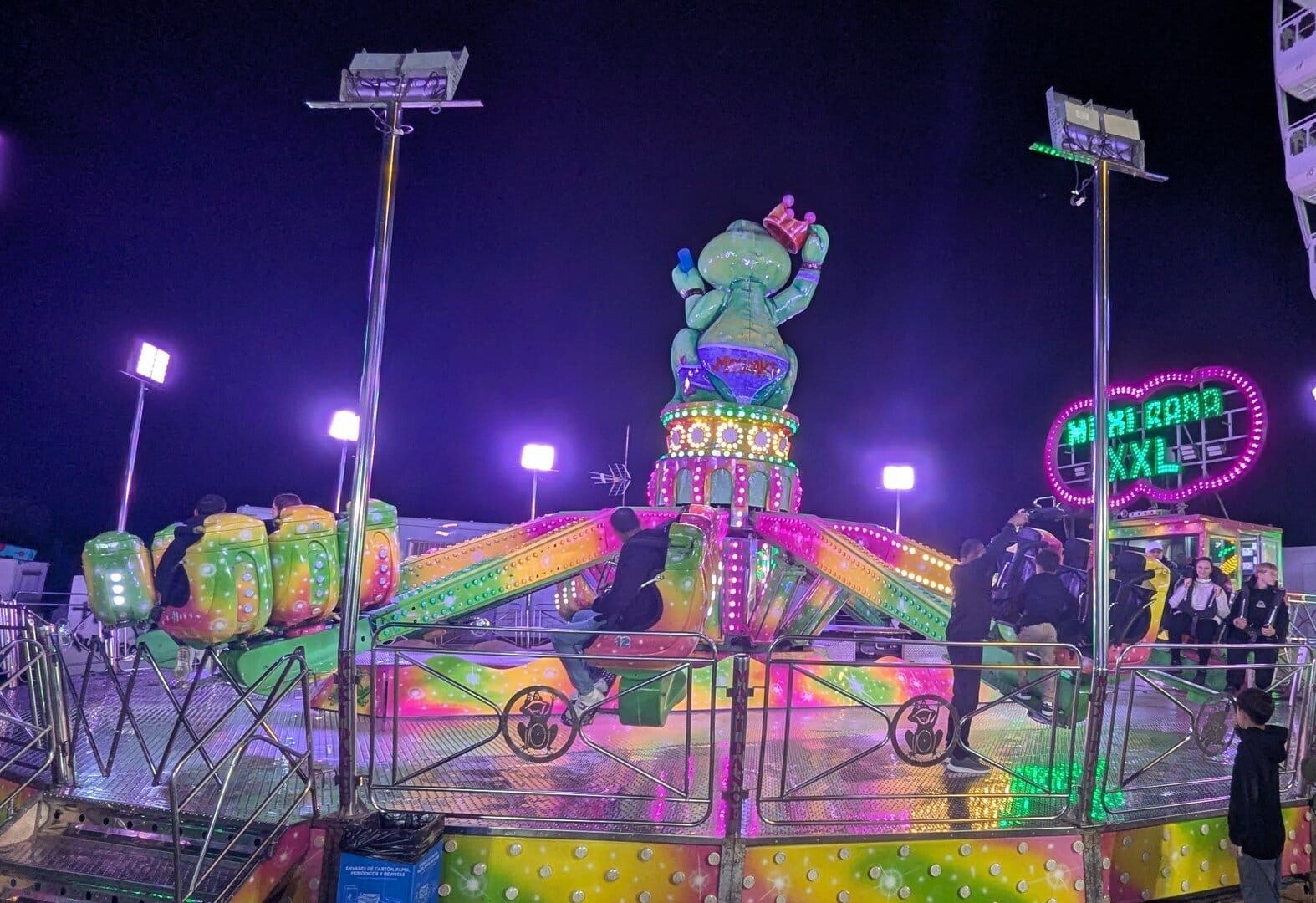 La atracción Maxi Rana XXL iluminada en verde y rosa, con la rana gigante coronada en lo alto y la noria al fondo