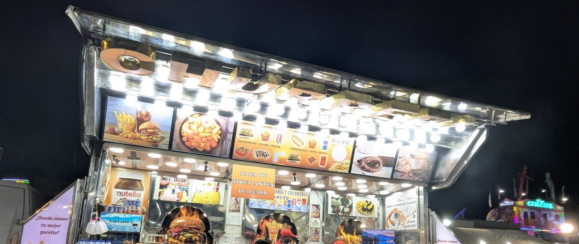 Churrería Canuto en la Feria de Todos los Santos de Dénia, con el menú iluminado y el cartel Saca tu tiquet antes de pedir