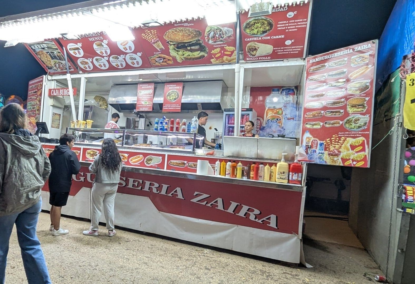Hamburguesería Zaira en la feria de Dénia, con el menú de kebab, cazuela con carne y rollo de pollo en carteles rojos